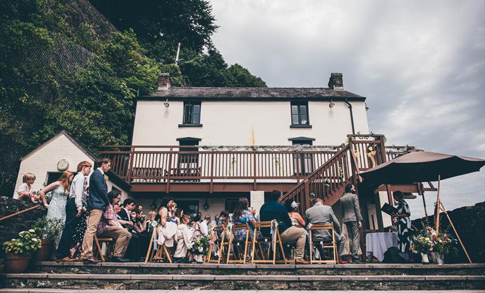 View of the back of a white-walled Boathouse looking up. A largely seated wedding party are on the patio in the foreground.